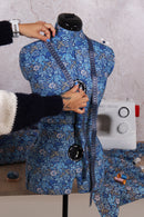 Person measuring a blue patterned garment on a mannequin with a sewing machine in the background.